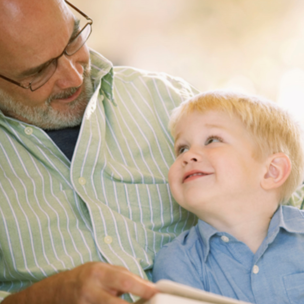 Young child sitting in grandfathers lap looking upward while reading a book following successful treatment from Little Allied Health