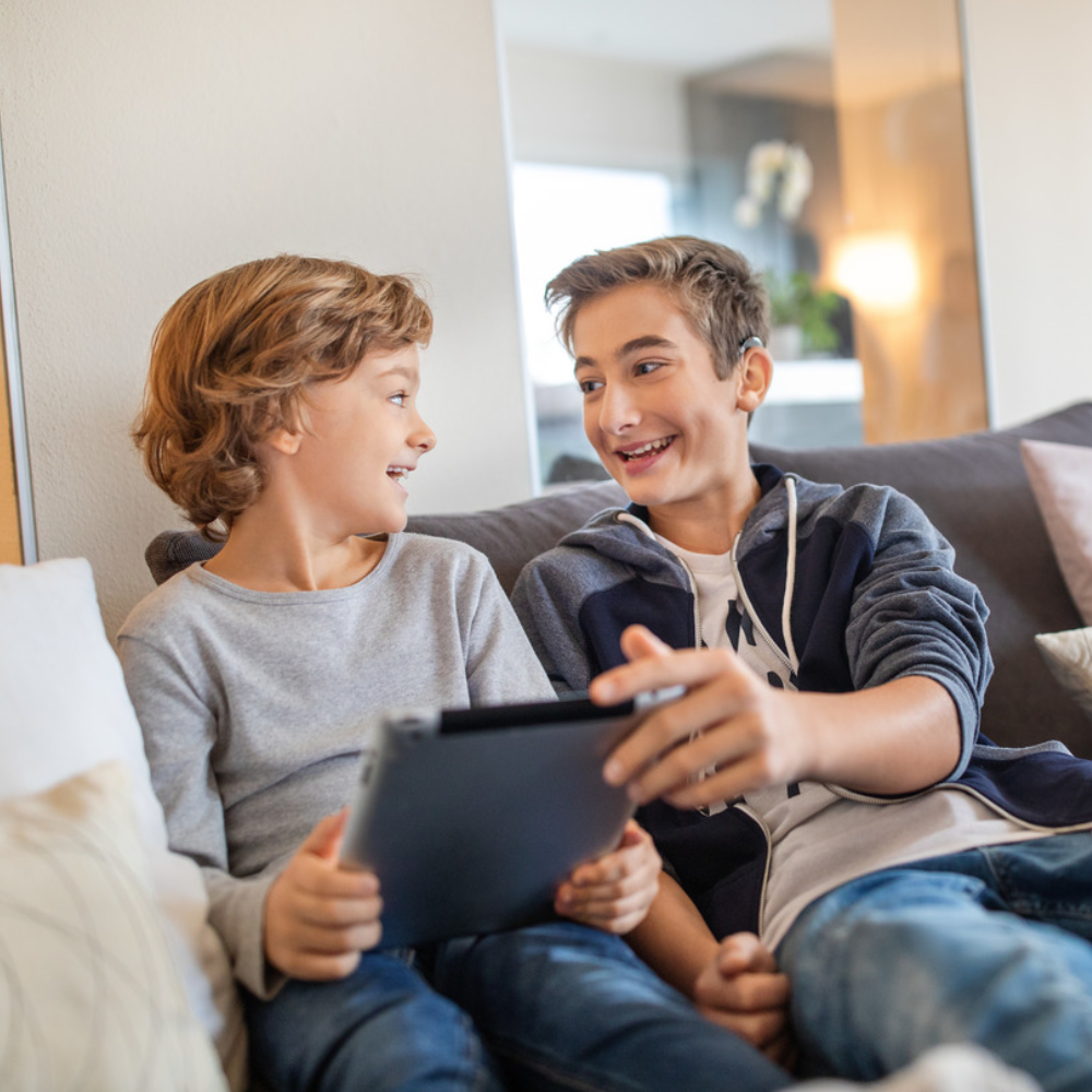 Teenagers sitting on couch using a mobile tablet with sound being transmitted via a MED-EL Synchrony Implantable Hearing Device